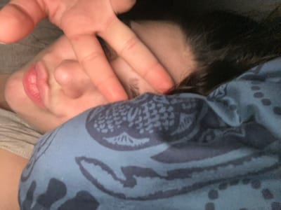 A person's face is partially visible under a blue patterned cloth. The face is tilted, with lips slightly pursed. A hand displaying a peace sign is positioned in front of the face, partially obscuring the nose and one eye. Dark hair is visible near the forehead. A gray garment is also partially visible. The overall impression is of someone playfully concealing their face.