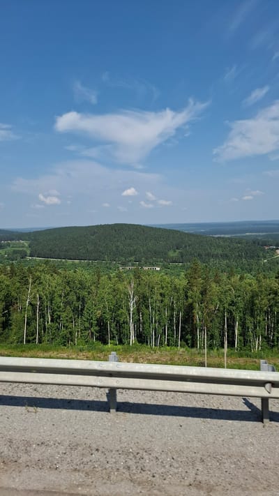 The image shows a scenic overlook with a guardrail in the foreground and a vast, verdant landscape stretching into the distance. The sky is a clear blue with scattered white clouds. The foreground consists of a gravel road and a silver metal guardrail. Behind the guardrail, a dense forest of green trees, including birches with white trunks, dominates the midground. A rounded, heavily forested hill rises in the background, with hints of a road winding around its base and some distant buildings or structures visible. The overall impression is one of natural beauty and tranquility, possibly a view from a roadside rest stop or a viewpoint in the region of Chistyye Klyuchi, Russia.