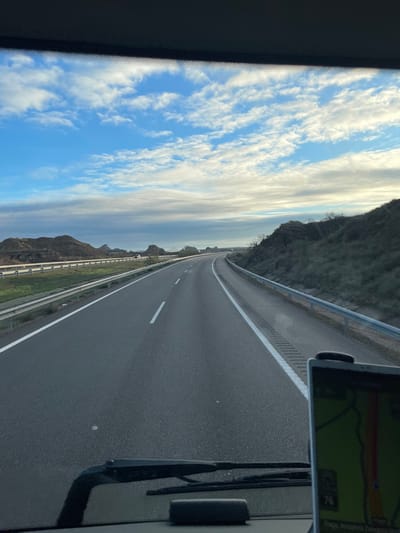 The image depicts a view from the interior of a vehicle, looking forward through the windshield onto a highway near Fraga, Spain. The sky occupies the upper half, featuring a blue expanse with scattered, white, streaky clouds. The highway is a multi-lane asphalt road, appearing to have two lanes in the direction of travel, marked with solid and dashed white lines. A textured rumble strip is visible along the right shoulder line. The road gently curves to the right in the distance.

On the left side of the highway, a metal guardrail separates the roadway from a lower terrain or adjacent road section. Beyond this, a landscape of undulating, arid, brown hills with sparse vegetation extends into the distance, characteristic of the Monegros region. A single faint vehicle is discernible on the left side of the highway in the mid-ground.

On the right side, another metal guardrail runs alongside a sloping embankment covered in dark green and brown scrub brush and sparse trees.

In the immediate foreground, the dark dashboard of the vehicle is visible, along with two windshield wipers in their resting position and a small black rectangular object resting on the dash. A GPS navigation device is mounted on the lower right of the windshield, displaying a map with a red route and a speed readout of "76." No people are visible.