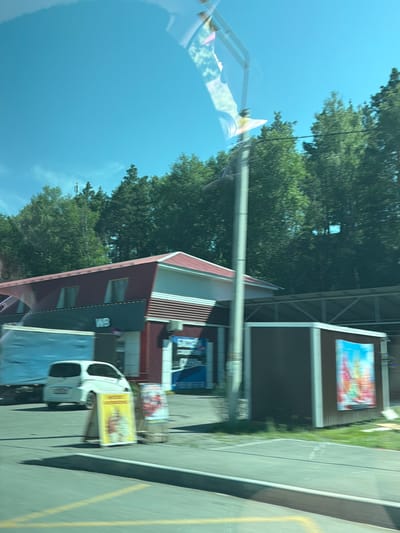 The image, taken from a moving vehicle, shows a commercial building with a red roof and white trim. A blue and white truck is parked to the left of the building, and a white car is parked in front of it. There are several signs displayed, including one with a yellow background and a picture of ice cream. The building appears to be a retail establishment. In the background, there are numerous trees and a clear blue sky. The image has a slight blue tint, possibly due to the lighting conditions or a filter. The location is identified as Uznezya, Russia.