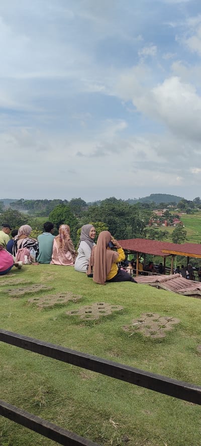 The image shows a group of people seated on a grassy hill overlooking a valley in Kecamatan Trawas, Indonesia. The coordinates provided are: -7.656 latitude and 112.594 longitude. The sky is partially cloudy. The group of people consists of seven individuals, four women and three men, seated on the hill. Some of the women are wearing traditional head coverings. The men are wearing shirts and pants. In the foreground, the ground is covered in short grass and a wooden railing. In the background, there is a view of the valley, with trees, houses, and a red-roofed building that appears to be a gazebo or restaurant. There are also people seated at tables under the gazebo. The scene appears to be peaceful and relaxing.