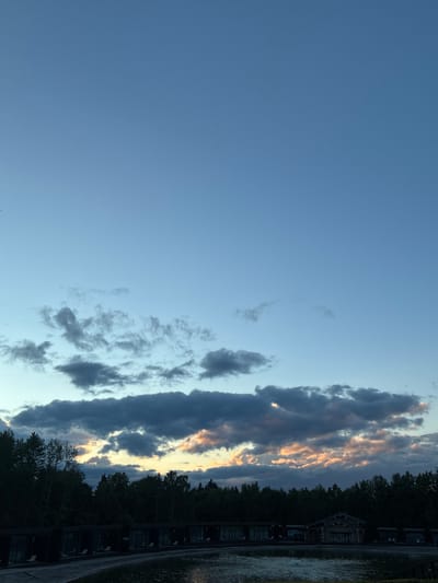 This is a media file of a sunset sky over a wooded area with some buildings and water. The sun is setting on the right side of the frame, casting a warm glow on the clouds. The sky is a pale blue, with streaks of orange and yellow from the setting sun. The clouds are dark and ominous, with patches of light breaking through.

On the right side of the frame, there are trees and buildings that appear to be part of a resort or a village. The buildings are dark and appear to be made of wood, with small windows. There is a body of water to the right of the buildings, reflecting the colors of the sky. The overall mood of the image is peaceful and serene, with a touch of melancholy.

The time of day is sunset, as indicated by the colors of the sky and the low angle of the sun. The weather is cloudy, but the clouds are not so thick as to obscure the sun completely. The location appears to be a natural setting, with trees and water, possibly a lake or a river. There are no people or animals visible in the image. There is no visible text in the image.

The image depicts a beautiful sunset over a serene landscape. The colors of the sky are vibrant, and the clouds add a dramatic effect. The buildings and trees create a sense of depth and perspective, while the water adds a reflective element. The overall impression is one of tranquility and natural beauty.