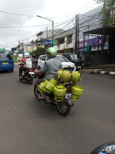 An urban street scene in Indonesia is captured under overcast skies. In the foreground, a male individual wears a light green helmet, a light grey sleeveless vest over a striped shirt, and dark shorts, riding a black motorcycle. This motorcycle is heavily laden with nine light green Pertamina 3kg LPG gas cylinders; six are secured on the rear rack, and three are visible on the left side. The motorcycle displays an Indonesian license plate, B 2795 MN. Ahead of this motorcycle, another motorcycle carries two individuals: the driver in a black helmet and jacket, and a passenger in a white helmet, red top, and a white backpack with an "AC MILAN" logo.
To the left, a blue van is partially visible. On the right side of the paved road, a white car with an Indonesian license plate (B 1383 PNR) is parked or moving slowly, followed by a black micro-van (Indonesian license plate B 1381 AGD). The right side of the street is lined with commercial buildings. One building prominently displays "XL SMART" and "Center" signage, referring to XL Axiata, an Indonesian telecommunications company, and another building features a green "X Center" sign. A person wearing an orange garment is seated on the sidewalk near these buildings. Overhead utility wires are densely strung across poles along the street.