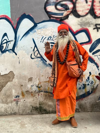 A man dressed in orange robes and a red cap stands in front of a graffiti-covered wall in Pushkar, India. He has a long white beard and is wearing glasses and a mala necklace. A small orange bag is slung over his shoulder. His left hand is raised, palm facing outwards. The wall behind him is weathered and peeling, with large graffiti tags in blue, white, and red. The ground in front of the wall is concrete, with some scattered debris.