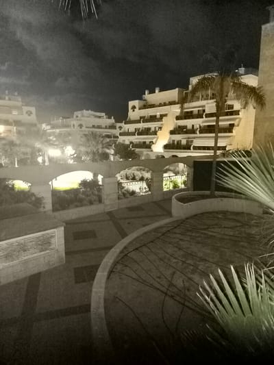 The image depicts a nighttime scene in Ashkelon, Israel, featuring a multi-story resort complex with several buildings. The architecture is modern, characterized by white facades, numerous balconies with railings, and illuminated windows. A palm tree stands prominently in front of one of the buildings, its fronds partially obscuring the structure.
In the foreground, a paved pathway with a geometric design leads into the complex. A curved planter bed with dried vegetation is visible in the lower right quadrant, and a large, spiky plant with green fronds occupies the far right edge of the frame.
A low stone wall with arched openings and a decorative metal fence runs horizontally across the middle ground, separating the pathway from a landscaped area. This landscaped section is illuminated by bright lights, highlighting lush green foliage. Beyond this, more resort buildings are visible, receding into the background.
The sky above is dark and cloudy, with faint streaks of light suggesting distant fireworks or a light effect. The overall impression is that of a festive or celebratory evening at a resort.
