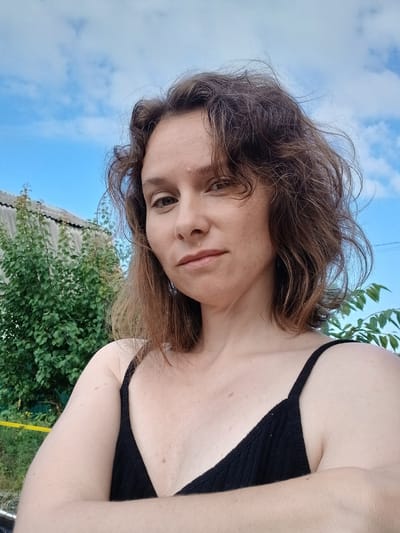 A woman with shoulder-length brown hair is pictured in a selfie taken outdoors. She is wearing a black tank top and has her arms crossed. Behind her, a green tree and a grey, corrugated metal roof are visible against a blue sky with scattered clouds. The environment appears to be rural, possibly in Maslova Pristan', Russia, with the presence of a residential building and vegetation.
