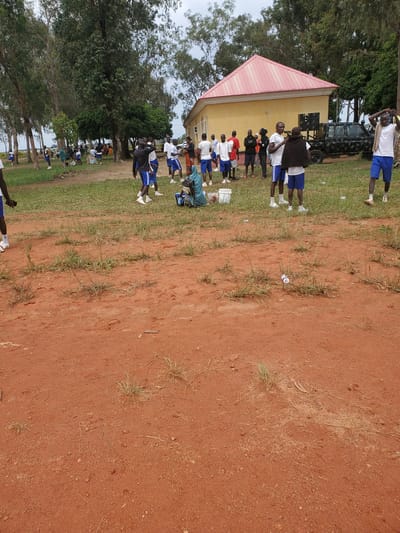 In Bahit, Nigeria, an outdoor scene unfolds with a group of young men in white t-shirts and blue shorts gathered near a yellow building with a red roof. A camouflage-patterned military vehicle is parked to the right of the building. Some individuals are standing, while others are seated or moving around on the grass and red earth. A woman in a blue headscarf is seated on the ground to the far left. The environment is characterized by sparse vegetation and tall trees in the background. The overall impression is that of a casual gathering or event.