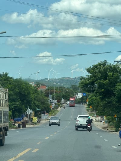 The image depicts a road scene in Lao Bảo Commune, Vietnam, characterized by a wide asphalt road with yellow and white lane markings. In the foreground, a white pickup truck with a license plate reading "74C-030 16" is traveling away from the camera, followed by a motorcycle with a rider wearing a blue helmet. To the left, a grey SUV is seen moving in the opposite direction, its license plate partially visible. Further up the road, a red semi-truck is also heading away from the viewer. The roadside is lined with lush green trees and vegetation, with some buildings and signs visible in the distance. The background features rolling hills dotted with numerous white wind turbines, indicating a renewable energy installation. The sky is a bright blue with scattered white clouds, and power lines are strung across the upper portion of the frame. The scene appears to be a typical day with moderate traffic.