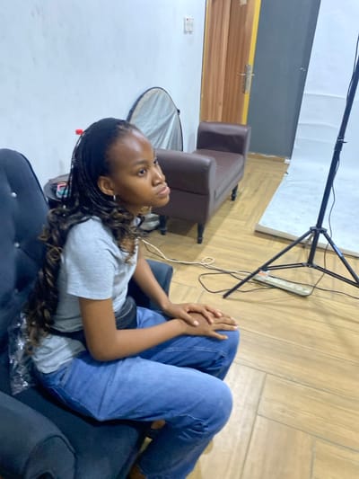 A young, dark-skinned woman with long braided hair sits relaxed in a dark-colored armchair, wearing a gray t-shirt and blue jeans. She gazes upward with a serene expression, her hands clasped on her lap. The setting appears to be a makeshift photography studio, evident from the light stand and reflector disk positioned nearby. In the background, there's a brown loveseat, a white backdrop, and a wooden door, giving the impression of an indoor, possibly residential, space.