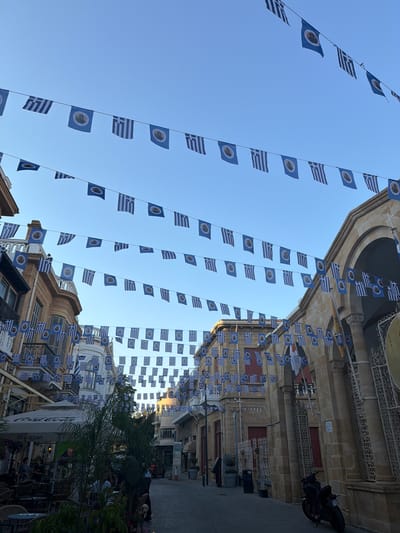 The image depicts a street lined with buildings adorned with numerous Greek flags strung across the sky. The flags alternate between the Greek national flag (blue and white stripes with a white cross on a blue canton) and a circular emblem on a blue background. These flags are hung on wires, creating a festive or commemorative atmosphere.

In the foreground, cafe tables and chairs are visible, suggesting an outdoor dining area. Some people are seated at the tables, appearing to be patrons of the cafe. Lush green foliage, including palm trees, adds to the setting. In the background, the architectural style of the buildings suggests a historical or traditional urban environment. A motorcycle is parked near one of the buildings.

The time of day appears to be late afternoon or early evening, with the sunlight casting a warm glow and creating some shadows. The sky is clear blue, indicating fair weather. The presence of the flags strongly suggests a national celebration or commemoration related to Greece or Cyprus, given the historical and cultural ties between the two countries. While no explicit text is legible on the flags, the overall scene conveys a sense of cultural pride and community gathering. The location is identified as Nicosia, Cyprus.