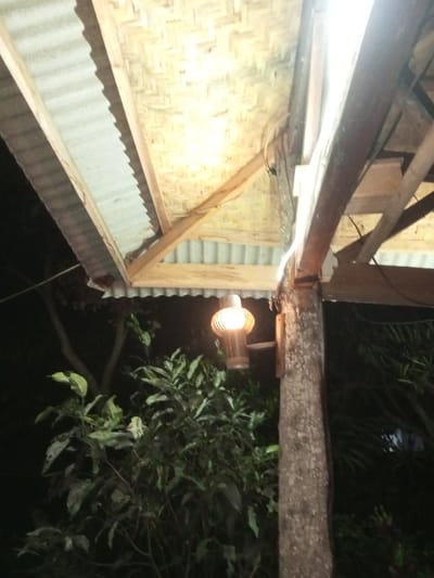 This image depicts a nighttime outdoor or semi-outdoor scene in Naya andura, India. The upper portion of the frame shows a roof structure, partially covered with corrugated metal sheeting and partially with woven bamboo or thatch panels, supported by visible wooden beams. A thick, textured tree trunk serves as a vertical support column on the right side of the frame. Attached to this tree trunk is a single, spherical lantern-style light fixture with a visible bulb inside, emitting a warm, bright glow. Electrical wiring runs along the roof structure and down the tree trunk, connecting to the light fixture. The background, below the roof and to the left of the tree, consists of dense, dark green foliage and tree branches, indicating surrounding vegetation in low light conditions. No people or specific actions are discernible.