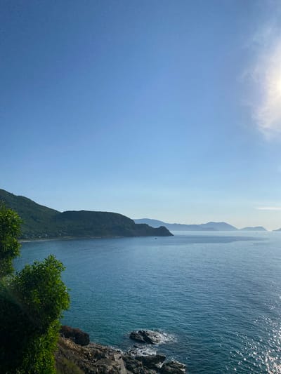 This image captures a serene coastal landscape in Cam Lam, Vietnam, under a clear blue sky. The foreground features lush green foliage on the left, with rocky terrain sloping down towards the sea. The ocean, a vibrant turquoise, stretches towards the horizon, its surface glinting with sunlight. In the mid-ground, a range of verdant, tree-covered mountains forms a dramatic backdrop along the coastline. Small structures, possibly houses, are visible nestled at the base of the mountains. Further out, a series of hazy, layered mountains fade into the distance. A few small boats are dotted on the calm water. The bright sun is positioned in the upper right corner, casting a warm glow and a slight haze in the sky. There are no people or animals visible in the scene.
