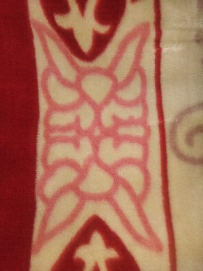 The image is a close-up, high-angle shot of a fuzzy, textured blanket with a pattern. The left side of the blanket is a solid, deep red. To the right of the red band, there is a cream-colored section with a repeating pink and red stylized floral or geometric pattern. The pattern is symmetrical and appears to be woven or printed onto the fabric. The texture of the fabric is evident, with visible fuzziness and light reflecting off the fibers, giving it a slightly glittery appearance in some areas. No people, actions, or specific environmental features beyond the blanket itself are visible. There is no information in the image to suggest a location in Haveli Jhangranwali, Pakistan.