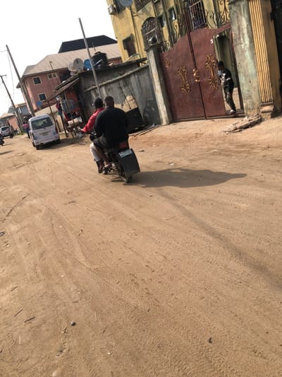 A street scene with a sandy road. A person rides a scooter with a package on the back. Buildings line one side of the road, including one with a brown gate. A vehicle is parked on the side of the road near the buildings. A shadow stretches across the road.