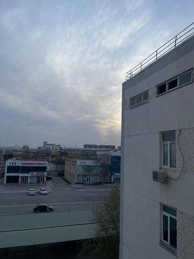 This image captures an elevated view of a street and commercial buildings in Navoiy City, Uzbekistan, under an overcast sky with a subtle sunbreak on the horizon. The perspective is from a multi-story building, a section of which occupies the right side of the frame. This building is light-colored with a textured facade, featuring multiple rectangular windows with white frames and an exterior "SHIVAKI" brand air conditioning unit mounted below a lower window. Its flat roof, topped with a dark metal railing, is visible against the sky.

Below, a two-lane asphalt road runs horizontally. A dark-colored sedan is positioned in the nearer lane. Across the road, two white sedans are parked parallel to the curb. Lining the far side of the street are commercial establishments. To the left, a two-story building features a dark glass ground floor and lighter upper levels. Signage on this building reads "LAZER MED CENTER," "LOR LABORATORIYA MARKAZI," and "SHAMS DORIXONA" (Shams Pharmacy). A dark vehicle is partially visible behind this structure. Adjacent to it, another two-story building displays "DIAGNOSTIK SENTR" and "LOR" signage, along with "NEVROPATOLOG," "TERAPEVT," and "UROLOG." Deciduous trees with sparse green foliage are scattered throughout the scene, and a large, dark green corrugated metal roof covers a structure in the immediate lower foreground. The distant background reveals a cityscape with various buildings, including taller structures and a discernible construction crane.
