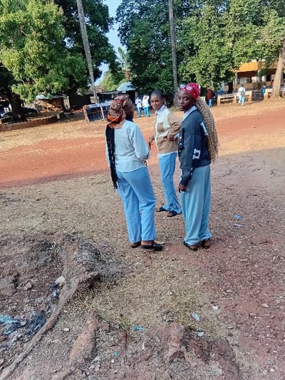 Three individuals stand on a dirt road near some trees. The person to the right wears a light-colored long-sleeved shirt and blue pants. The middle person wears a white and brown sweater with blue pants. The person on the left wears a black and white jacket with blue pants. All three are wearing shoes. A wooded area and building structures are in the background. The ground is covered with dirt and dry grass.