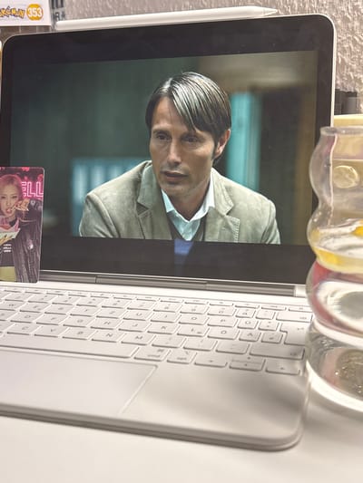 A light-colored laptop is positioned on a white surface, displaying a close-up of a man's face on its screen. The man, with graying hair and a neutral expression, wears a light brown blazer over a light blue collared shirt. To the left of the laptop screen, a rectangular trading card featuring a young woman with pink hair is placed upright. To the right of the laptop, a clear glass object filled with a light yellow liquid and possibly ice or fruit slices is visible. The laptop's keyboard is in focus in the foreground. There is no identifiable information to suggest a location in Velbert, Germany.
