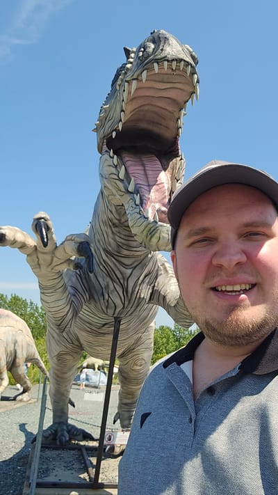 A man wearing a grey baseball cap and a grey and black polo shirt is taking a selfie with a large Tyrannosaurus Rex statue. The T-Rex statue is light grey with darker grey stripes and has its mouth open, revealing pink inside and sharp white teeth. Its left arm is raised. The man is smiling and facing the camera. In the background, there is a clear blue sky, some green trees, and other dinosaur statues, including what appears to be a Brontosaurus. There are also signs and some gravel on the ground. The scene appears to be an outdoor attraction, possibly a dinosaur park, located in Saint-Léonard-d'Aston, Canada.