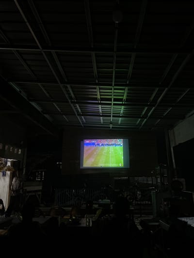 The media file shows an interior view of a space, possibly a covered outdoor area or a large room with a corrugated metal ceiling and support beams. A projection screen is illuminated, displaying a soccer match with player statistics and commentary. The scene is dimly lit, suggesting it is either evening or nighttime. The angle of the image is unusual, with the top of the frame appearing at the bottom, implying the camera was held upside down or the image has been rotated incorrectly.

On the left side of the image, the corrugated metal wall and structural framework dominate the scene, creating a repetitive pattern. A circular white object, possibly a light fixture or a sensor, is visible on the wall. The projection screen, positioned on the right side, is the focal point of the illuminated area, showing a vibrant green soccer field with players. Text is visible on the screen, including what appears to be a score and player information.

The right side of the image transitions into a darker area, revealing indistinct objects that could be furniture, such as chairs, and possibly some outdoor elements like railings or a gate. The overall impression is one of a casual viewing environment, perhaps a gathering of people to watch the soccer game, though no people are clearly visible. The lighting, the content on the screen, and the structure of the space suggest a relaxed atmosphere. The location context provided is Khang Tanjong, Indonesia, which doesn't offer specific visual cues within the image itself.