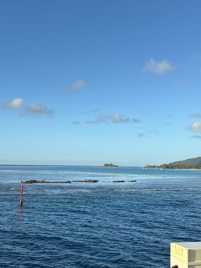 This image depicts an expansive maritime scene in Fa'a'ā, French Polynesia, under a clear blue sky with scattered cumulus clouds. The foreground features rippled blue ocean water, with a red cylindrical navigation buoy positioned vertically on the left. Across the mid-ground, several dark, irregular reef formations or submerged rocks are visible just at or below the water's surface. In the distant center, a small, densely vegetated island (motu) is present. To the far right, a verdant coastline of a larger island with green hills extends, along which approximately five to seven sailboats are anchored. A light-colored, rectangular structure, partially visible in the bottom right corner, suggests the vantage point from which the photograph was taken. No individuals are explicitly visible, though the sailboats imply human presence in the broader environment.