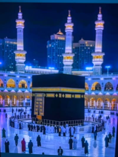 The image depicts the Kaaba in Mecca, Saudi Arabia, at night. The structure is a large, black cuboid with a golden embroidered band around its upper portion. It is surrounded by a plaza where numerous people in traditional Islamic attire are walking and gathered. In the background, there are tall, illuminated minarets and modern buildings, suggesting the presence of a city surrounding the holy site. The scene is lit with artificial lights that cast a blue and purple hue over the environment. The image does not depict Cadek, Indonesia.