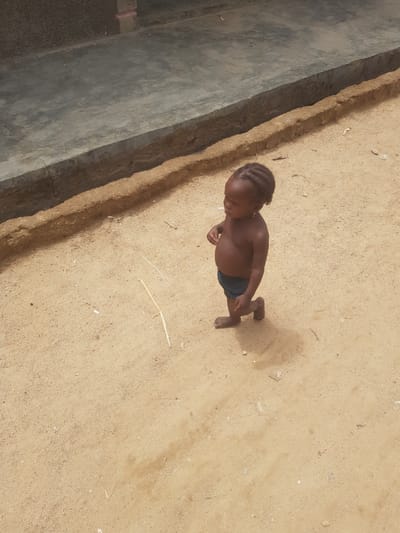 A young child stands barefoot on a sandy surface next to a concrete structure. The child has dark skin, braided hair, and is wearing dark-colored shorts. The child is facing towards the concrete structure. The sand is light brown and covers the ground. A few small pieces of debris are scattered on the sand. The concrete structure is gray and appears to be a wall or curb. The location appears to be outdoors in Dutsinma B, Nigeria.