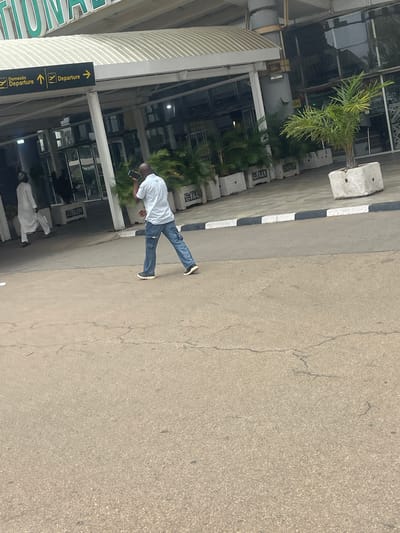 The image, captured in Gwaska, Nigeria, presents an outdoor scene in front of an airport terminal under bright daylight. The main subject is a man wearing a white collared shirt, blue jeans, and dark shoes, holding a smartphone. He is walking on a paved area, seemingly a pedestrian path, with the airport building to his left.
Further back, closer to the building, another person in white traditional attire is visible. The modern airport terminal features extensive glass facades and a distinctive curved, ribbed canopy. Lush green palm trees and other tropical plants are positioned in front of the building, enhancing the landscaping.
A black and white striped curb delineates the paved area from an asphalt road, which shows minor cracks. Along this edge, several concrete barriers are placed, some bearing partially legible text that appears to be "SAAN" or "SARAN."
Prominent directional signs are visible near the building, indicating routes. These black signs with yellow text and arrows read "Departure" multiple times, and one specifically states "Domestic Departure." A large green sign, partially visible on the building's facade, likely spells out "INTERNATIONAL" or the airport's name. The scene suggests a clear, sunny day at a functional airport.