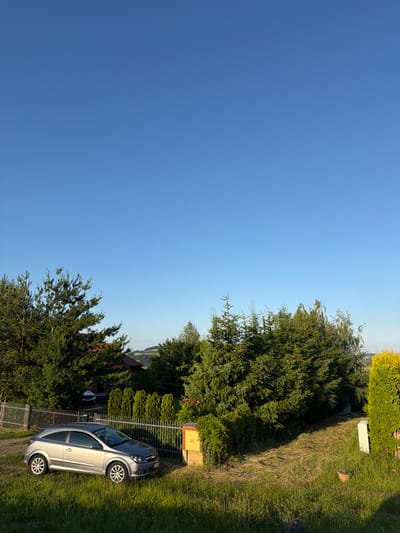 The image depicts a residential scene with a silver hatchback car parked on a grassy area next to a metal fence. In the background, there are trees, a house with a red roof, and rolling hills under a clear blue sky. A yellow sign with Polish text is visible on a brick structure. The overall setting suggests a rural or suburban environment, possibly in Sienna, Poland, given the context.