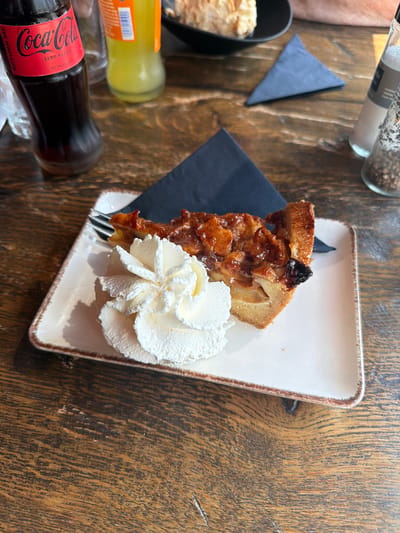 A slice of apple pie with whipped cream sits on a white plate with a brown rim. A fork is partially visible, resting on the plate. The pie is topped with a glossy glaze and appears to contain chunks of apple and possibly some dark dried fruit. The whipped cream is artfully piped into a swirl. In the background, a dark bottle of Coca-Cola Zero Sugar is visible, along with a clear bottle of what appears to be a yellow beverage. A dark grey folded napkin is placed behind the pie. The scene is set on a wooden table with a rustic grain. The lighting suggests an indoor setting, possibly a cafe or restaurant. The image was captured in Hilversum, Netherlands.
