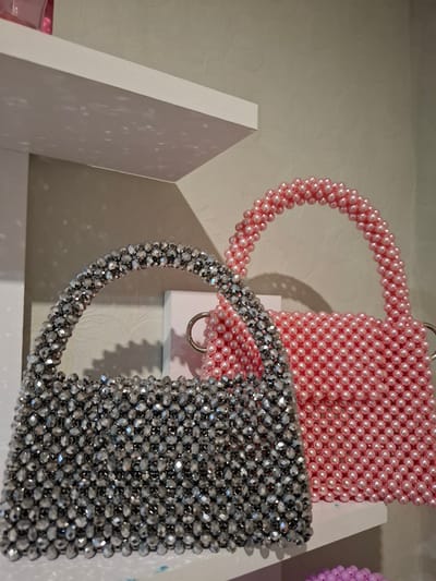 The image displays two beaded handbags on a white shelf. The handbag on the left is covered in faceted, silver-colored beads, while the handbag on the right is covered in round, pale pink pearls. Both bags have top handles made of the same beaded material. A portion of a pink glass object is visible on the top shelf in the upper left corner. The background is a textured, light-colored wall. The scene is indoors, with soft lighting creating shadows on the wall. The location is identified as Korenovsk, Russia.