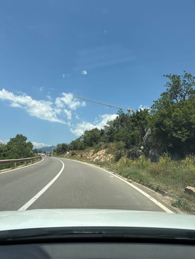 The image captures a view from a vehicle driving on a winding road in Drobnići, Montenegro. The road curves to the left, with a white line marking its center and right edge. On the left side of the road, there's a metal guardrail and lush green vegetation. The right side of the road is bordered by rocky terrain and dense green trees. In the distance, several cars are visible on the road, and a large construction crane extends horizontally across the sky above the trees. The sky is bright blue with scattered white clouds. The lower portion of the image shows the hood of the white vehicle from which the photo is taken.