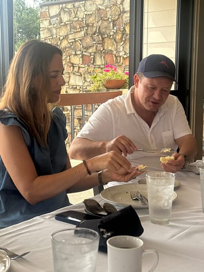 The image displays two individuals, one female and one male, seated at a white-tablecloth-covered table in what appears to be a restaurant or café. The specific location within Montgomery, United States, cannot be determined from the visual information provided.
The female subject, positioned on the left, has long brown hair and wears a light blue, short-sleeved top with ruffled shoulders. Her left hand holds a small piece of light-colored bread or pastry with a fork, suggesting she is applying a spread. A thin bracelet and a dark-banded watch or bracelet are visible on her left wrist. Her gaze is directed downwards towards her food.
The male subject, on the right, wears a white short-sleeved collared shirt and a navy blue baseball cap featuring a red marlin logo. A light-colored chain is visible around his neck. His left hand holds a piece of light-colored bread or pastry, and his right hand holds a silver knife, which he appears to be using to spread a substance on the bread. His gaze is also directed downwards.
The table setting includes two white plates, assorted silver cutlery (forks and knives), clear glasses filled with ice water, and a white ceramic mug in the foreground. Additionally, a dark-colored smartphone, sunglasses, and a folded black napkin displaying a white Under Armour logo are present on the table.
The background, visible through a large window or open section of the dining area, reveals an outdoor setting. A stone wall constructed from irregularly shaped, multi-toned stones (tan, gray, and reddish hues) is prominent. A wooden railing runs horizontally along the lower portion of the wall. Beyond the railing, potted plants with vibrant pink flowers and green foliage are visible. Green trees are present in the distant background under a bright sky, indicating daytime.