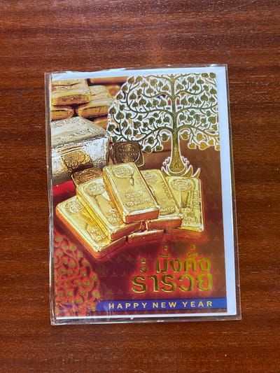 This media file is a close-up overhead shot of a New Year's greeting card. The card is placed on a wooden surface.
The card features a striking design with a prominent illustration of stacked gold and silver bars. The gold bars appear to be the primary focus, with some having visible markings. To the left of the bars, there is an intricate golden design resembling a tree with heart-shaped leaves. The background of the card is a rich, deep red, adorned with gold floral patterns. On the right side of the card, a blue vertical strip contains the text "HAPPY NEW YEAR" in white, sans-serif font. Above the blue strip, in a stylized golden font, are some characters that appear to be in a script other than Latin, likely Thai given the location context.
The card is encased in clear plastic, suggesting it might be a newly purchased item or one intended for mailing. The lighting is even, indicating it was likely taken indoors during the day. No people are visible in the image. The overall impression is one of prosperity and celebration, aligning with the theme of a New Year's greeting.