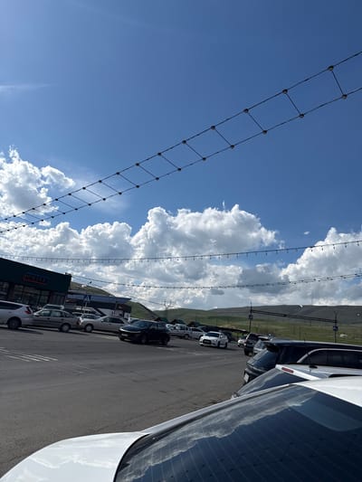 The image shows an outdoor scene in Tsovagyugh, Armenia. A parking lot with multiple parked cars is visible along a paved road. Behind the parking lot, a grassy hillside stretches towards the horizon. Overhead, the sky is mostly blue with fluffy, white cumulus clouds. Two types of overhead lighting structures are visible. One is a simple string of lights. The second is a grid of cables with evenly spaced dark connectors. The lighting structures are suspended above the parking lot. A building with the name "UNIPACK" can be seen. The image is oriented sideways.