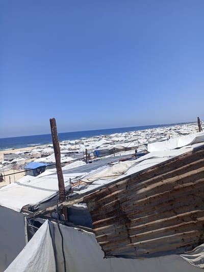 The image depicts a large refugee camp situated near a coastline under a clear blue sky. Numerous white tents, likely makeshift shelters, are densely packed together, stretching as far as the eye can see towards the sea. A wide expanse of calm blue water is visible beyond the camp, meeting the horizon. The sand of the beach is visible between the tents and leading down to the water.
In the foreground, a rusted, corrugated metal structure, possibly a damaged roof or wall, partially obscures the view of the tents. A dark, weathered metal pole stands prominently in the center of the frame, also appearing to be part of the temporary structures. Some tents are made of white tarp material, while others appear to be constructed with different fabrics, including a blue tent visible in the middle ground. The overall impression is one of a crowded and precarious living situation.
The scene suggests a bright, sunny day with no visible clouds. The location is identified as Gaza, though the specific site within Gaza is unknown. No specific activity or event is clearly discernible, but the sheer number of tents indicates a significant population of displaced people. The image conveys a sense of scale and the challenging conditions faced by those living in the camp. There is no visible text in the image.
