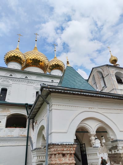 This is a low-angle, vertical medium shot of the golden domes of a Russian Orthodox church, likely the Church of the Resurrection in Faliukovo, Russia, under a cloudy sky. The church features white walls, ornate architectural details, and multiple gold-plated onion domes topped with crosses. A smaller, green-tiled roofed structure is visible in the foreground, obscuring part of the church's facade. A person wearing a camouflage shirt is visible in the lower right corner of the image, facing away from the camera. The overall impression is one of grandeur and religious significance.