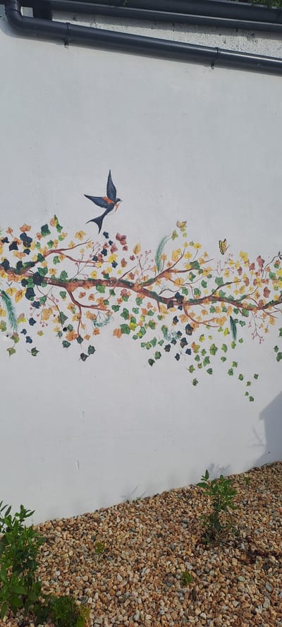 The image shows a white wall with a mural of a tree branch with falling leaves. A bird is depicted in flight above the branch, and a butterfly is visible to the right. Below the wall, there is a ground covered in small, light-colored pebbles. The scene appears to be outdoors, with natural light casting shadows. There are no people or identifiable structures visible in the immediate vicinity that would indicate a specific location within Castlebar, Ireland. The mural is the primary subject, rendered in a stylized, artistic manner.