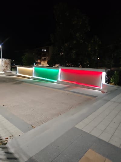 This is a nighttime, eye-level, medium shot of an outdoor public space in Peshtera, Bulgaria, featuring a modern illuminated art installation. The installation is a horizontal, rectangular structure divided into three sections, each glowing with a different color: yellow on the left, green in the middle, and red on the right, resembling the Italian flag. The structure is set against a dark background with trees and a building visible. The foreground consists of paved walkways and a patterned stone surface. No people are visible in the frame. The overall scene is illuminated by the art installation and a street lamp on the left, creating a somber yet vibrant atmosphere.