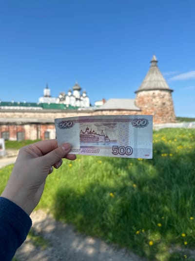 A hand holds a 500-ruble banknote in front of a blurred background. The background features a portion of the Solovetskiy Monastery complex in Solovetskiy, Russia. The monastery includes several whitewashed buildings with domed roofs and a round, stone tower with a conical roof. Green grass with scattered yellow flowers covers the ground in the foreground and midground. The sky is clear and blue.