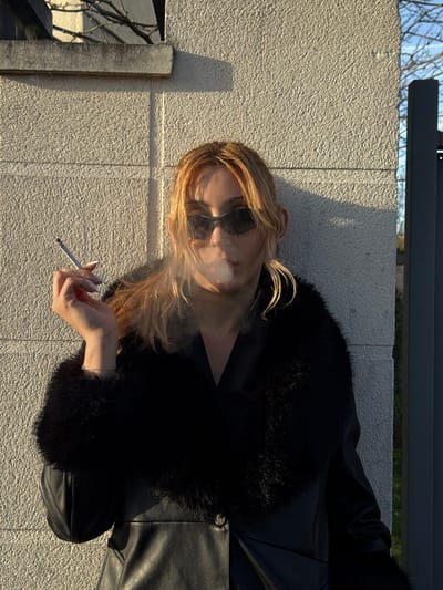 In Asnières-sur-Seine, France, a light-skinned individual, appearing female, stands outdoors during daylight. She wears dark wraparound sunglasses and a black coat with a fur-trimmed collar and cuffs. Her light-colored hair is styled to frame her face. She holds a lit cigarette between the index and middle fingers of her right hand, which displays white-tipped nails, and exhales a plume of smoke directly forward. The individual faces the viewer. Behind her is a textured, light-colored wall with vertical seams, illuminated by strong sunlight that casts a sharp shadow in the upper left background. Beyond the wall, bare tree branches are visible, and to the right, a dark gray vertical structure, possibly part of a gate or fence, is partially visible.