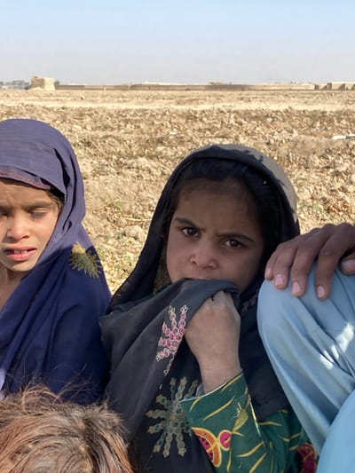 Mandisar, Afghanistan. The foreground depicts three individuals in an arid, outdoor environment. On the left, a young person, likely female, with light brown skin, wears a dark blue headscarf and a matching garment with a yellow sun-like embroidery. Her eyes are partially closed or downcast, and her mouth is slightly open. Centered, a young girl with darker skin wears a dark headscarf and a green garment with multi-colored patterns. Her right hand is raised, holding a fold of her headscarf to her mouth, and her brown eyes are directed towards the viewer. To her right, a portion of an adult male's arm and a leg in light blue trousers are visible. His hand rests on his leg. The immediate environment is characterized by dry, light brown, rocky soil with minimal vegetation. In the background, a long, low, light-colored wall or structure extends horizontally, with indistinct buildings and what appear to be solar panels visible further in the distance under a pale blue sky.