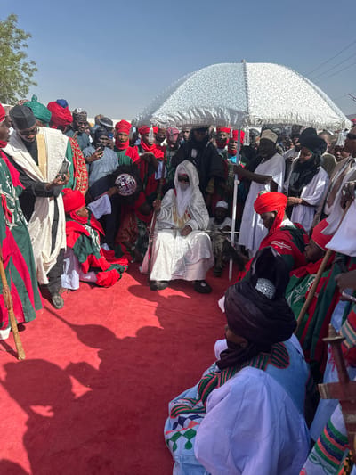 The image depicts a crowded outdoor ceremonial gathering in Zango, Nigeria. The foreground and middle ground are covered with a red carpet. Under a clear blue sky, a central figure is seated directly on the red carpet, wearing a white flowing robe, a white head covering that partially obscures the face, dark sunglasses, and dark shoes. This individual holds a long, ornate staff or scepter. A large, ornate, light-colored umbrella with a textured pattern is held above this central figure by a standing man in a white tunic.

Surrounding the central figure are numerous individuals, many seated or kneeling on the red carpet, and a dense crowd standing behind them. People are dressed in various traditional attire, predominantly consisting of colorful flowing robes and elaborate turbans or head coverings in shades of red, green, white, black, and blue.

To the left of the central figure, a person in a white and green striped robe and a dark head covering is visible, holding a smartphone. Directly in front of the central figure, a person in red and green robes kneels, holding what appears to be a ceremonial cap or turban towards the seated figure. Other individuals seated on the carpet also wear colorful robes and headwear, and some hold staffs. Strong sunlight casts distinct shadows onto the red carpet.