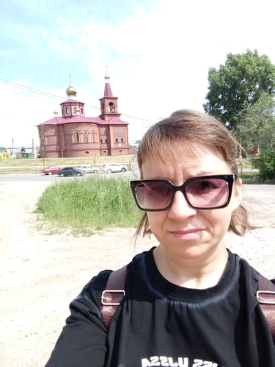 A woman in her 30s or 40s with short brown hair and sunglasses is taking a selfie outdoors. She is wearing a black t-shirt with a brown strap visible over her shoulder. She appears to be in a rural or semi-rural setting.

In the background, a red brick church with golden domes and a bell tower is visible. A road runs in front of the church, with several cars parked or driving on it. To the left of the church, there are some small houses and buildings. Tall green grass and trees surround the scene.

The weather appears to be sunny with some clouds in the bright blue sky, suggesting it is daytime, likely late morning or afternoon. The overall mood seems casual and relaxed. The woman's expression is neutral.

There is some text visible on the woman's t-shirt, though it is partially obscured and appears to be upside down. It reads "ASSLU ZZI".

The location is identified as Pavlovsk, Russia.