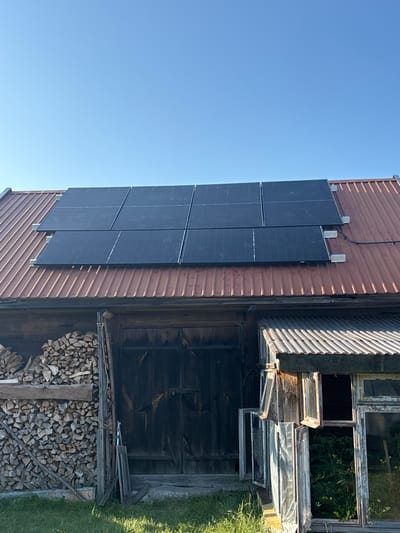 The image shows the exterior of a building with a corrugated metal roof, possibly a barn or shed, located in Janów, Poland. The roof is partially covered with a grid of dark solar panels. Below the roofline, there are wooden walls and large, dark wooden doors. To the left of the doors, a substantial stack of cut firewood is visible. To the right, there is a section of the building with a corrugated metal roof extension and multiple broken or open windows, some of which reveal greenery inside. The sky above is clear and blue. The ground in front of the building is covered with grass.