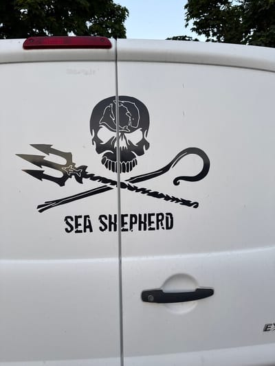 The image shows the rear of a white van parked outdoors in Saint-Orens-de-Gameville, France. A black decal is affixed to the van's rear doors, featuring a skull and crossbones design with a trident and a coiled rope, and the words "SEA SHEPHERD" below. The van is parked near a tree line, and the sky is visible above. A red brake light is mounted on the top of the van. The van's rear door handle is black and is visible in the lower right portion of the image. The van appears to be a commercial vehicle.