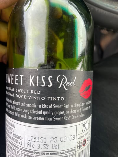 A close-up shot shows a green wine bottle with a black and white label. The label reads "SWEET KISS Red," "NATURAL SWEET RED," and "NATURAL DOCE VINHO TINTO" in large, stylized font. A red lipstick print is visible above the "SWEET KISS" text. Smaller text describes the wine as "sophisticated, elegant and smooth" and suitable for sharing with friends. The label also displays "750ml," a NAFDAC registration number, and an "18+" age restriction symbol. Information at the bottom of the label indicates that the wine is produced in South Africa for Gravel Junction Wine and Spirits, located on Road R44 in Klapmuts, Paarl, South Africa. It is further noted that the wine is imported/distributed by Lamtup Nigeria Limited, Plot 1, Opeyemi Bamidele Crescent, Phase 1, Lagos, Nigeria. A human hand is partially visible at the bottom of the frame. The background is dark and out of focus, suggesting the bottle is inside a vehicle or in a dimly lit area.