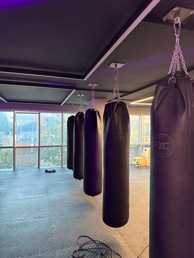 A row of four black punching bags hang from metal chains attached to a dark wall. The punching bags are of varying lengths and are arranged vertically. The top bag is the longest and has a circular logo with text that appears to read "GBC GOLDEN BELT CLUB". The wall on the left is dark and features a recessed rectangular frame. On the right side of the image, a large window looks out onto an urban scene with buildings and trees. The floor is covered in grey interlocking puzzle mats. A coiled black cable lies on the floor near the window. The lighting suggests it is daytime, with natural light coming through the window. The setting appears to be a gym or boxing studio. There are no people visible in the image.