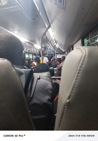 The image is taken inside a bus with passengers. The composition is framed between two headrests, focusing on the rows of people in the background. The interior lighting is soft, highlighting the metallic handrails and overhead advertisements. People are seated and standing, filling the space with varied clothing and postures. The overall impression is that of a typical commute within a public transport setting.