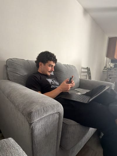 A man with dark, curly hair, a mustache, and a short beard sits comfortably on a large, grey fabric couch. He wears a black t-shirt with an indistinct graphic and black pants, along with an earring in his left ear. He holds a smartphone in both hands, looking down at its screen with a slight smile, suggesting amusement or contentment. An open black laptop rests on his lap, and some folded papers or money are visible next to it on the couch.
The grey couch features thick cushions and armrests. The background walls are plain and light-colored. To the right, a partially visible kitchen area with cabinets and appliances, and a light-colored chair or stool, suggests an open-plan living space. The casual indoor setting, likely a living room or common area, takes place in Redondo Beach, United States, depicting a moment of relaxation or casual engagement with technology. No legible text is visible in the image.