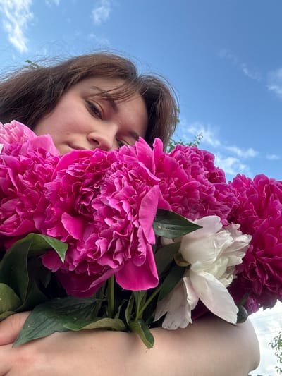A close-up, low-angle shot shows a young woman holding a large bouquet of pink and white peonies against a bright blue sky with scattered white clouds. The woman's head is tilted downwards, her dark brown hair falling around her face, which is partially obscured by the flowers. Her eyes are closed, and she appears to be looking down or resting her head on the bouquet. The peonies are vibrant and fully bloomed, filling most of the frame. The sky occupies the upper portion of the image, suggesting an outdoor setting. No specific location in Niva, Russia, can be determined from the visual information.
